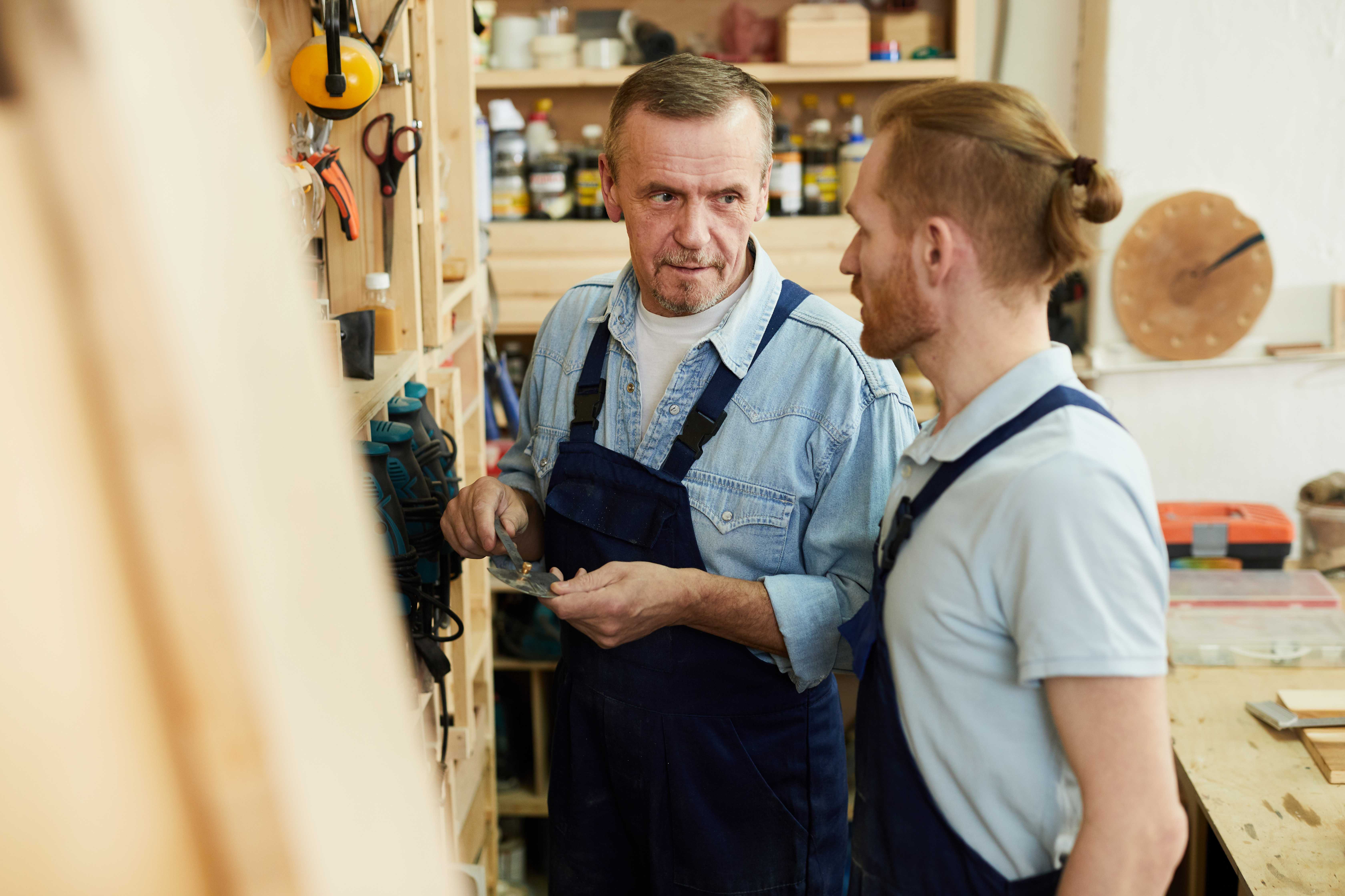Portrait of senior carpenter talking to young trainee while working in joinery workshop, copy space